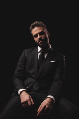 Takım elbiseli ciddi bir adam karanlık stüdyo arka planında gözlük tutarken poz veriyor.