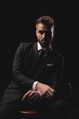 Takım elbiseli ciddi zarif bir adam karanlık arka planda otururken elinde gözlüklerini tutuyor.