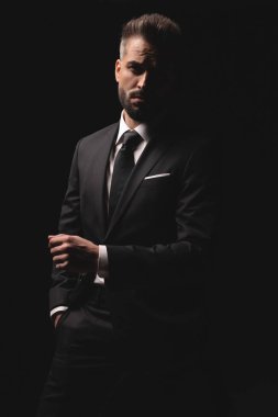 Siyah takım elbiseli ciddi bir işadamı, bir eli cebinde duruyor ve elini bir yumruğuyla siyah stüdyo arka planında tutuyor..