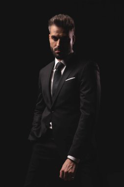 Takım elbiseli ciddi bir adam karanlık stüdyo arka planında bir eli cebinde duruyor.