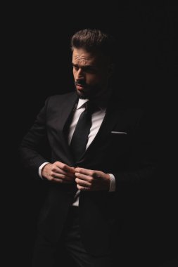 Takım elbiseli şık bir adam karanlık stüdyo geçmişine bakarken ceketinin düğmelerini ilikliyor.