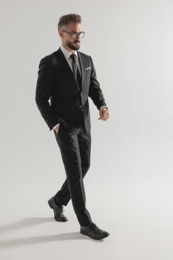 Siyah takım elbiseli ciddi bir iş adamının, gri stüdyo arka planında tek eli cebinde yürürken yan görüntüsü.