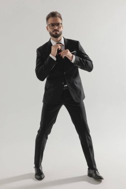 Takım elbiseli yakışıklı zarif adam gri stüdyo arka planında siyah kravatını düzeltirken ayakta duruyor.