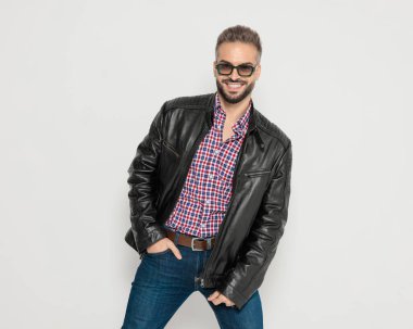 Güneş gözlüklü ve deri ceketli neşeli çekici adam gülümsüyor ve gri stüdyo arka planında el ele poz veriyor.