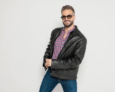 Güneş gözlüklü mutlu ve çekici adam gri arka planda poz verirken gülümsüyor ve deri ceketini tutuyor.