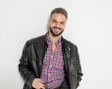 Deri ceketli mutlu adam, gri arka planda güneş gözlüğünü tutarken gülümsüyor.