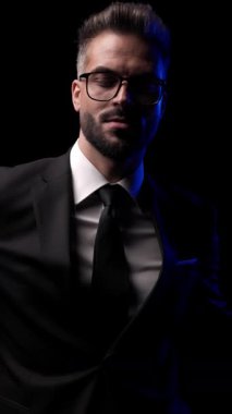 Takım elbiseli ve gözlüklü şık bir adam etrafa bakıyor, ceketinin düğmelerini ilikliyor, ve koyu arka planda mavi ışıkla yürüyor..