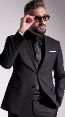 Güneş gözlüklü ciddi bir adamın ellerini ceplerine koyduğu ve kravatını gri arka planda düzelttiği dikey bir video..