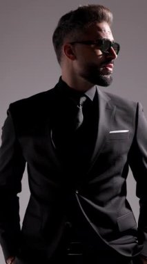 Güneş gözlüklü şık adam etrafa bakıyor, kravatını düzeltiyor, gözlüklerini ayarlıyor ve gri stüdyo arka planından uzaklaşıyor..