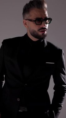 Güneş gözlüklü ciddi bir adam öne doğru yürüyor, ceketini tamir ediyor, ellerini ovuyor ve gri arka plana bakıyor..