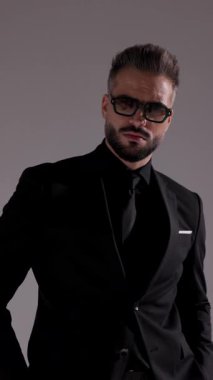 Güneş gözlüklü, kendine güvenen, gözlüklerini takan ve gri stüdyo arka planında yürüyüp giden bir adamın yakın çekimi..