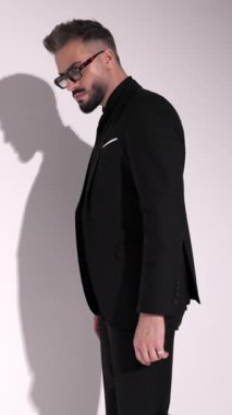 Siyah takım elbiseli ve güneş gözlüklü ciddi bir adam yürüyor, ceketini açıyor ve beyaz arka planda düğmelerini ilikliyor..