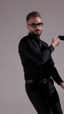 Güneş gözlüklü ve siyah elbiseli havalı adam ceketini omzuna geçirip gri arka planda yürüyüp gidiyor..