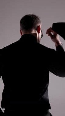 Güneş gözlüklü ve siyah elbiseli bir adam yürüyor, ceketini omzuna takıyor, gözlüklerini tamir ediyor ve gri arka planda ceketini tutuyor..