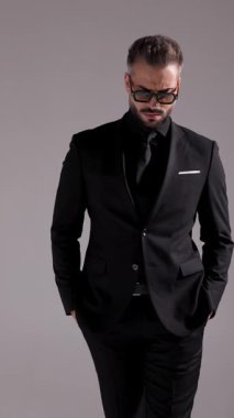 Güneş gözlüklü ve siyah takım elbiseli havalı adam elleri ceplerinde poz veriyor, etrafa bakıyor ve gri arka planda yürüyüp gidiyor..