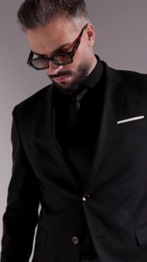 Güneş gözlüklü, siyah takım elbiseli, kollarını kavuşturup gri arka planda dudaklarına dokunan düşünceli bir adamın dikey videosu..