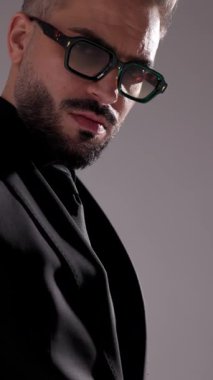 Güneş gözlüklü, siyah takım elbiseli, gri stüdyo geçmişine bakan ciddi bir adamın yan görüntüsü..