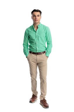Yeşil kareli gömlekli, elleri ceplerinde beyaz arka planda poz veren kendinden emin genç bir adam.