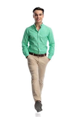 Yeşil kareli gömlek ve bej pantolon giyen mutlu genç adam, beyaz arka planda elleri ceplerinde ileri doğru yürüyor.