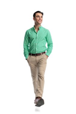 Neşeli genç adam, şık günlük kıyafetler giyer, elleri ceplerinde yürürken gülümser ve beyaz arka planda yan tarafa bakar.