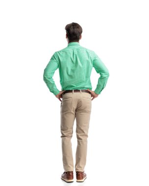 Yeşil kareli gömlek ve bej pantolon giyen, elleri beyaz arka planda bekleyen kendine güvenen genç bir adam.