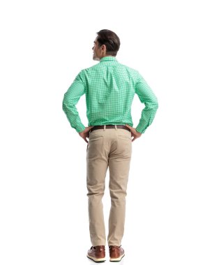 Yeşil kareli gömlek ve bej pantolon giyen, elleri kalçasında bekleyen ve beyaz arka plana bakan sıradan bir genç adam.