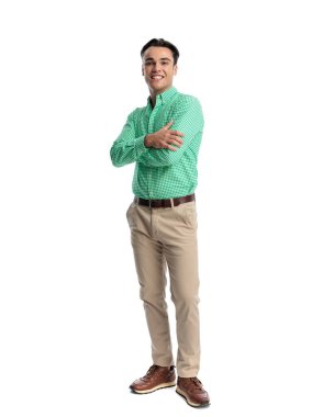 Yeşil gömlek ve bej pantolon giyen kendine güvenen genç adam beyaz arka planda ileriye bakarken kollarını kavuşturup gülümsüyor.