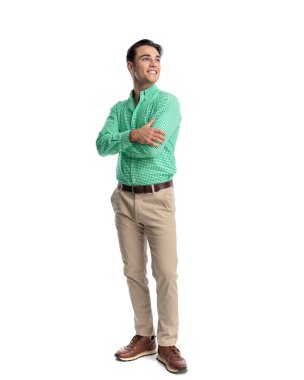Yeşil gömlekli ve bej pantolonlu neşeli genç adam kollarını çapraz yapıp beyaz arka plana bakıyor.