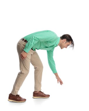 Meraklı bir adamın öne doğru eğildiğini ve elini yere doğru uzattığını yeşil ekose gömlek ve beyaz arka planda izole edilmiş bej pantolon giyerek bir şeyler almak için uzandığını gördüm.