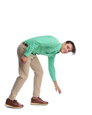 Neşeli, sıradan bir adam öne eğiliyor ve yan tarafa bakarken yere uzanıyor ve beyaz arka planda yeşil gömlek ve bej pantolon giyiyor.