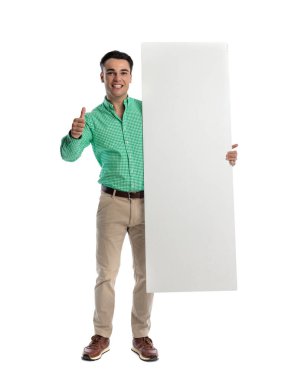 Neşeli bir adam elinde kocaman boş bir tahtayla ayakta dikilirken ve beyaz arka planda ileriye bakarken baş parmak işareti yapıyor.