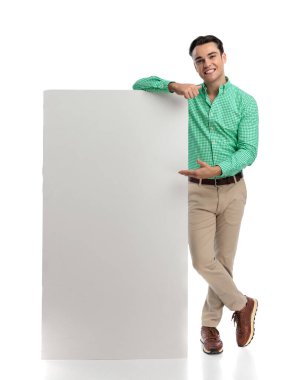 Dirseğini boş bir reklam panosuna yaslayıp önüne bakıp beyaz arka planda poz veren mutlu bir adam.