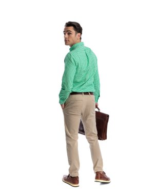 Yeşil kareli gömlek ve pantolon giyen şık bir adam deri evrak çantası tutarken beyaz arka plana bakmak için arkasını dönüyor.