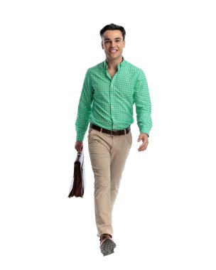 Yeşil kareli gömlek ve pantolon giyen mutlu genç adam kahverengi evrak çantasını tutarken ileri doğru yürüyor ve beyaz arka planda ileriye bakıyor.