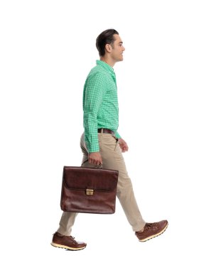 Yeşil kareli gömlek giyen ve beyaz arka planda yürürken deri evrak çantası tutan sıradan gülümseyen bir adam.