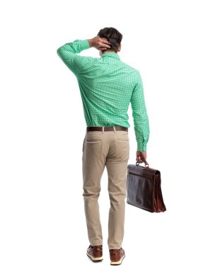 Kafası karışmış bir adam şık giyinmiş, beyaz arka planda izole edilmiş deri bir evrak çantası tutarken kafasını kaşıyor.