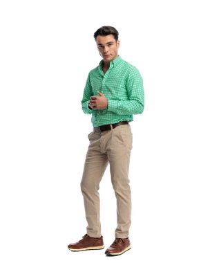 Yeşil kareli gömlek ve bej pantolon giyen kendine güvenen genç adam elleri bağlı, beyaz arka planda tek başına ileriye bakıyor.