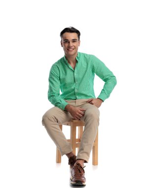 Yeşil kareli tişört ve bej pantolon giyen neşeli genç adam beyaz arka planda izole edilmiş, ileriye bakan ahşap taburede oturuyor.
