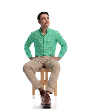 Umut dolu genç adam yeşil kareli gömlekli, tahta taburede oturuyor, elleri kalçasında, beyaz arka plana bakıyor.