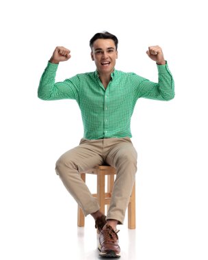 Heyecanlı genç adam yeşil kareli gömlekli tahta taburede oturuyor ve beyaz arka plana bakarken yumrukları havada tezahürat yapıyor.