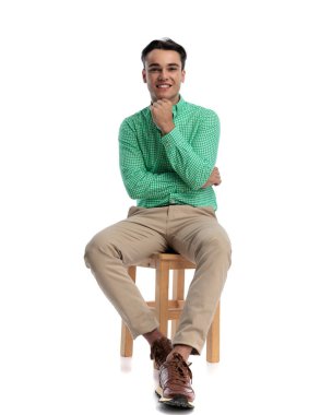 Tahta bir sandalyede oturan, çenesine dokunan ve beyaz arka planda kameraya gülümseyen kendinden emin genç bir adam.