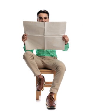 Ahşap bir sandalyede oturan, elinde kocaman boş bir gazete tutan ve beyaz arka planda ileriye bakan mutlu genç adam.