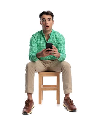 Ahşap bir taburede oturan genç adam elinde akıllı bir telefonla beyaz arka planda izole edilmiş afallamış bir ifadeyle ileriye bakıyor.