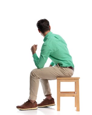 Ahşap bir taburede oturan ve beyaz arka plandaki ilgiyle geriye bakan meraklı genç bir erkek.