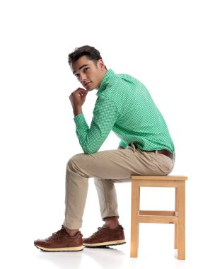 Tahta bir taburede oturan ve beyaz arka planda ileriye bakarken çenesine dokunan düşünceli bir erkeğin yan görüntüsü