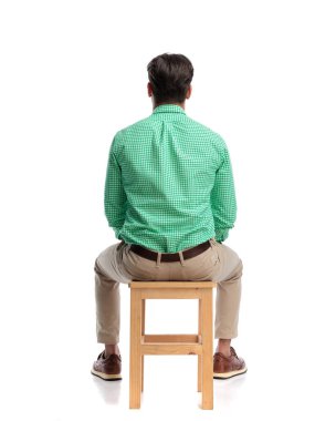 İşaretli gömlek giyen ve beyaz arka planda tahta taburede oturan sıradan bir adamın arka görünüşü.