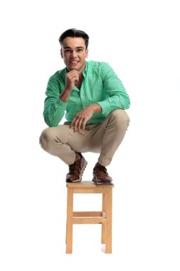 Uzun boylu, yeşil tişörtlü, tahta taburede çömelmiş beyaz arka planda ileriye bakarken çenesine dokunan neşeli bir adam portresi.