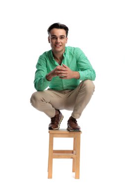 Ahşap tabureye çömelmiş, elleri sımsıkı bağlı, beyaz arka plana bakarken mutlu, sıradan bir adam.