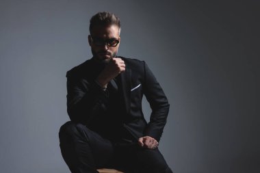 Siyah takım elbiseli dalgın moda adamı, koyu gri arka planda güneş gözlüğü takarken çenesine dokunuyor.