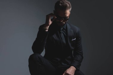 Siyah takım elbiseli havalı gizemli adam, koyu gri arka plana bakarken güneş gözlüğünü ayarlıyor.